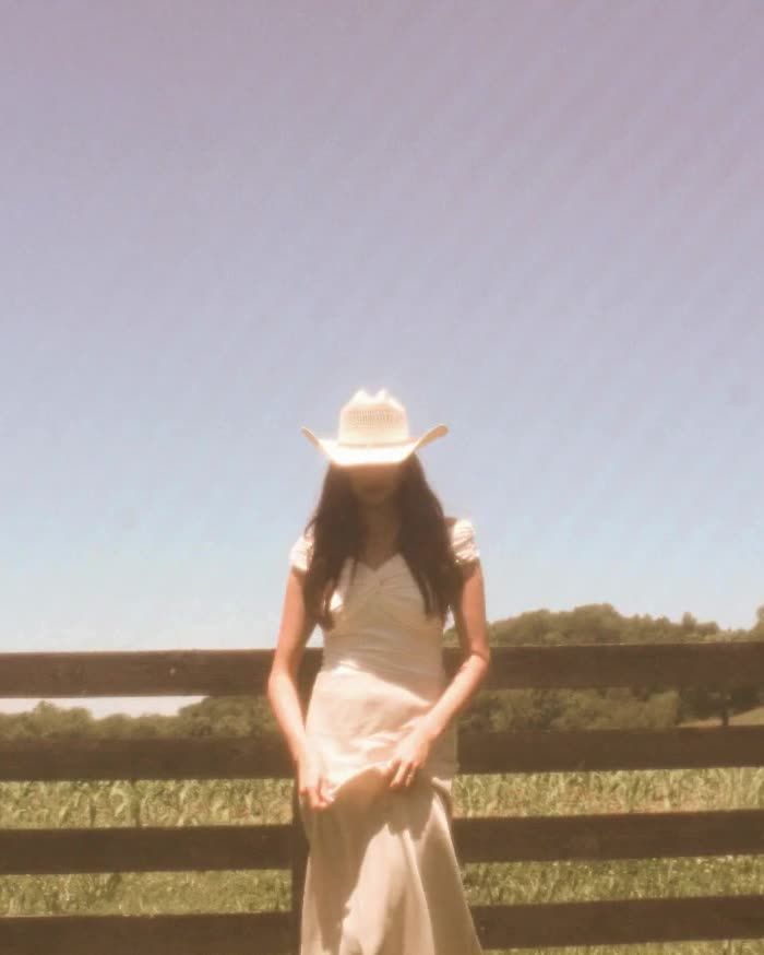 A woman in a white dress and cowboy hat at a fence, in warm film color